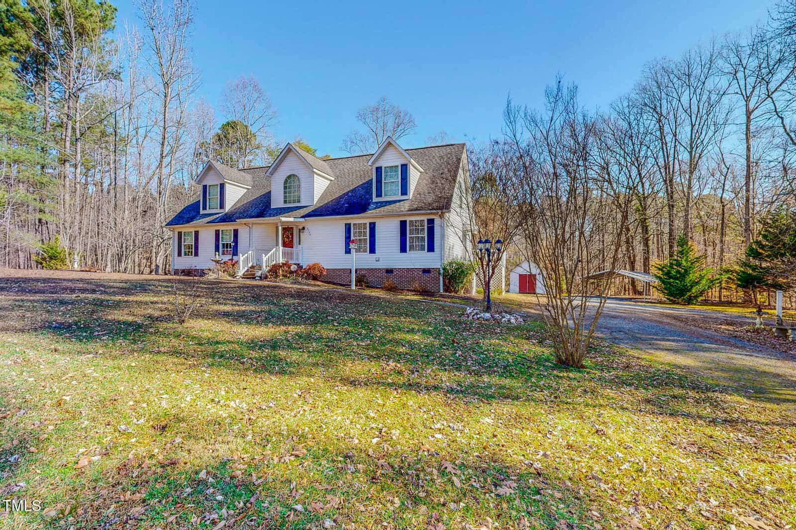 A two-story white house with a dark roof is surrounded by trees. The home features a front porch and multiple windows. A driveway leads to a detached garage on the right. The grassy yard is scattered with fallen leaves under a clear blue sky.