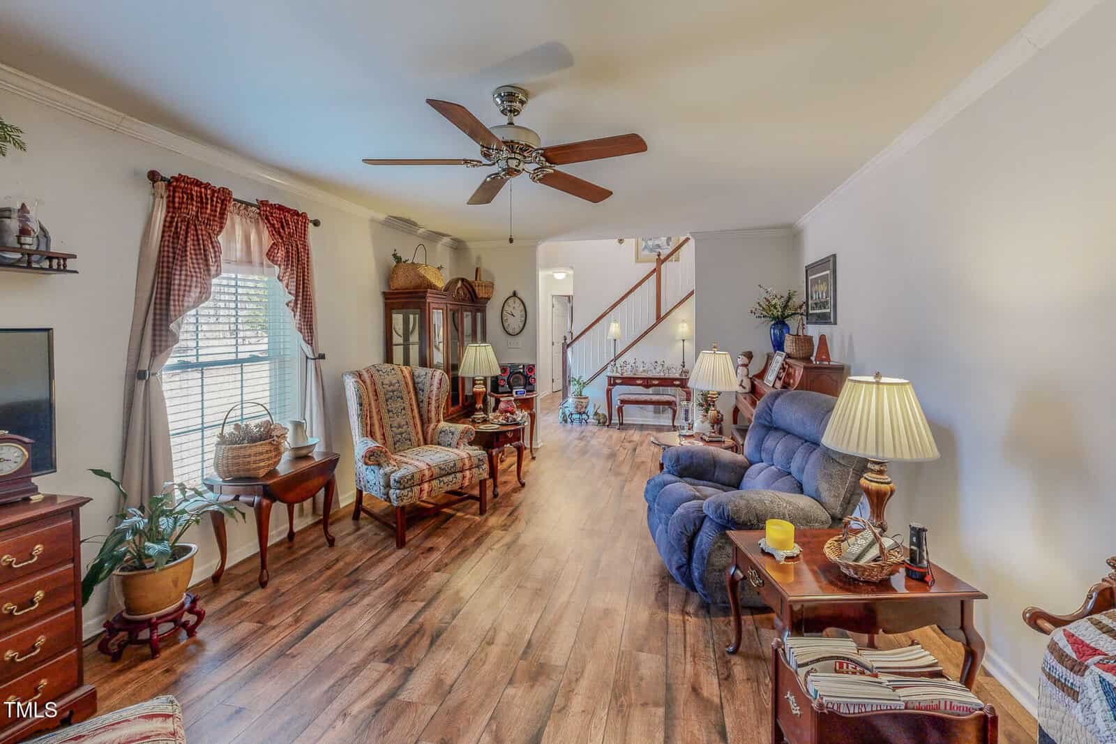 A cozy living room with hardwood floors, featuring two armchairs, a blue recliner, and multiple lamps. There's a ceiling fan, plants, and decorative items. A staircase is visible in the background, and sunlight streams through a window with plaid curtains.