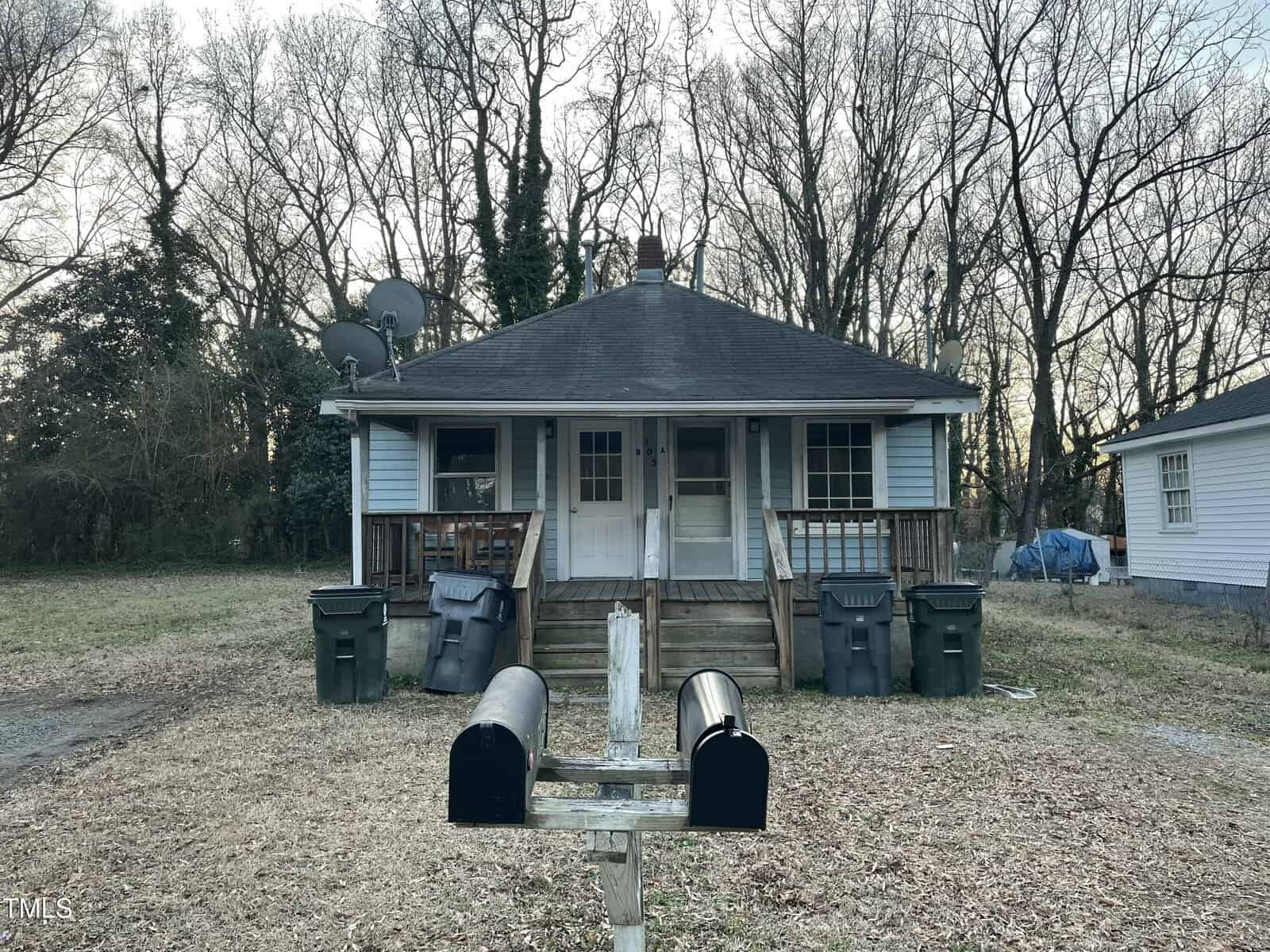 Nestled at 105 Hunt Street in Oxford, this quaint single-story house features a welcoming front porch with dual entrance doors. A yard of leafless trees surrounds it, while multiple trash bins line the steps. Two black mailboxes stand in the foreground, and antennas perch on the roof.