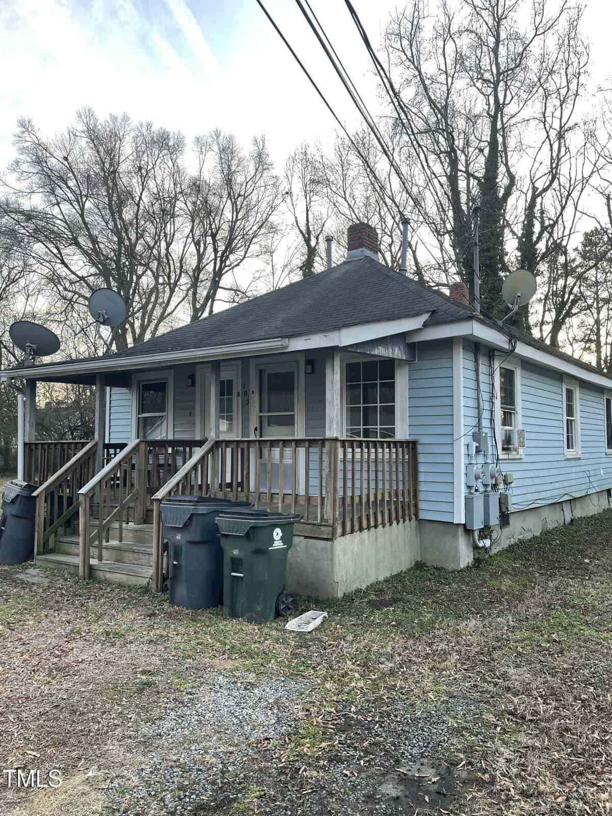 A small blue house with a dark roof has a front porch featuring wooden railings and a set of stairs. There are three satellite dishes attached to the house. Trees without leaves surround the property, and trash bins are near the porch.