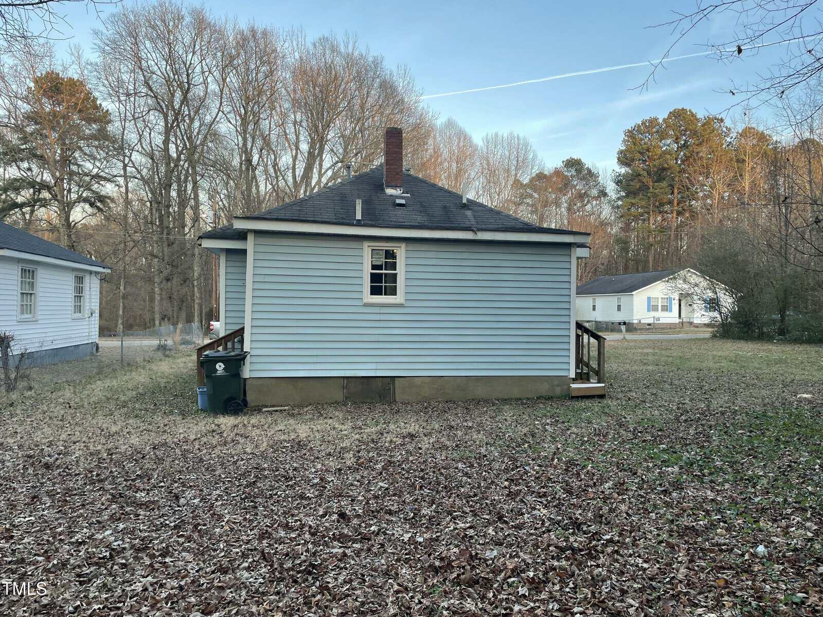 A small, single-story house with light blue siding and a dark roof is shown. There is a chimney, one visible window, and a few steps leading to a side door. The house is surrounded by leaf-covered ground, with trees and another house in the background.
