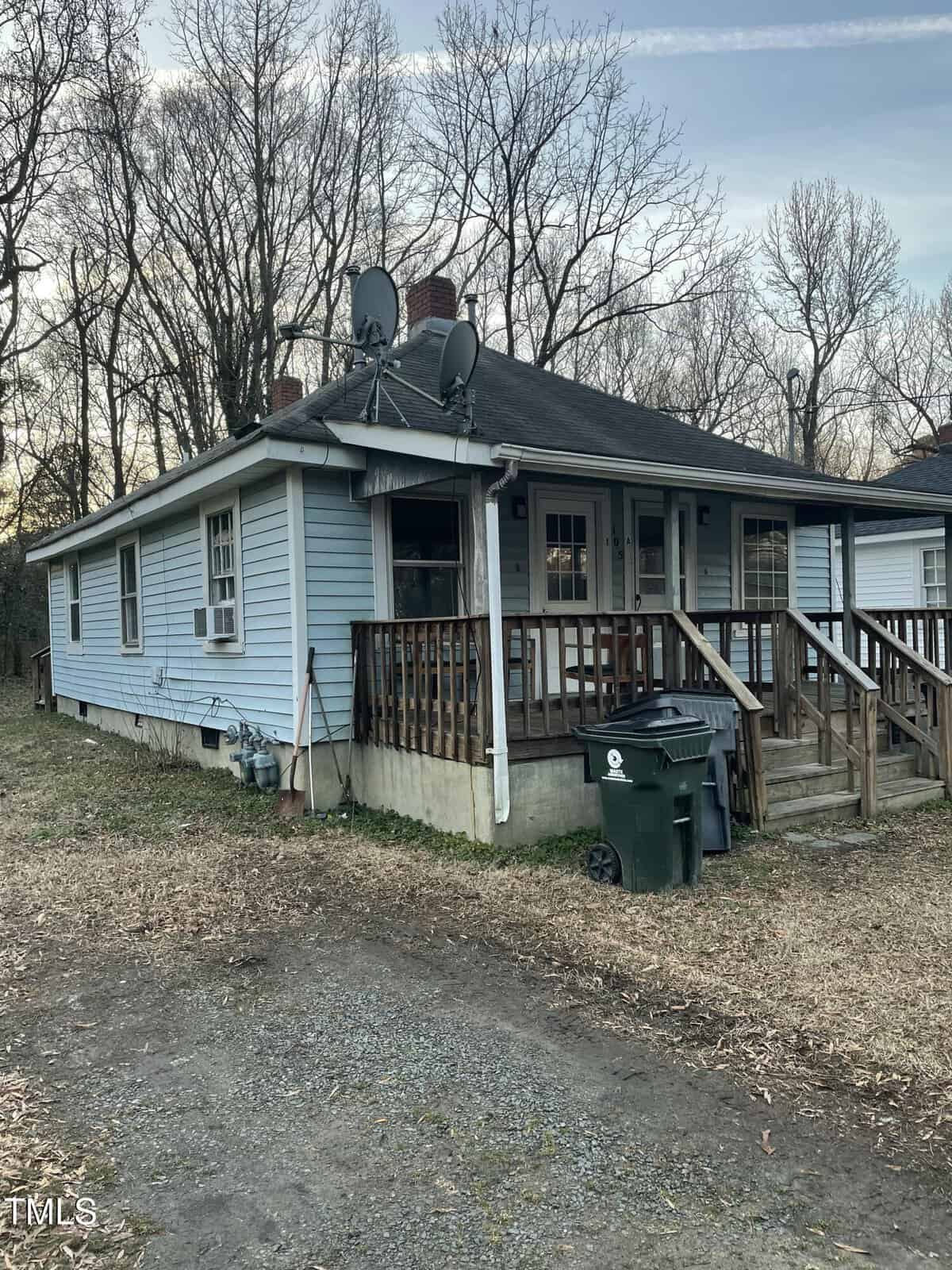 A small blue house with a covered porch, two satellite dishes on the roof, and a wooden railing around the porch. There are trash bins near the entrance, and the house is surrounded by bare trees and a patchy lawn.
