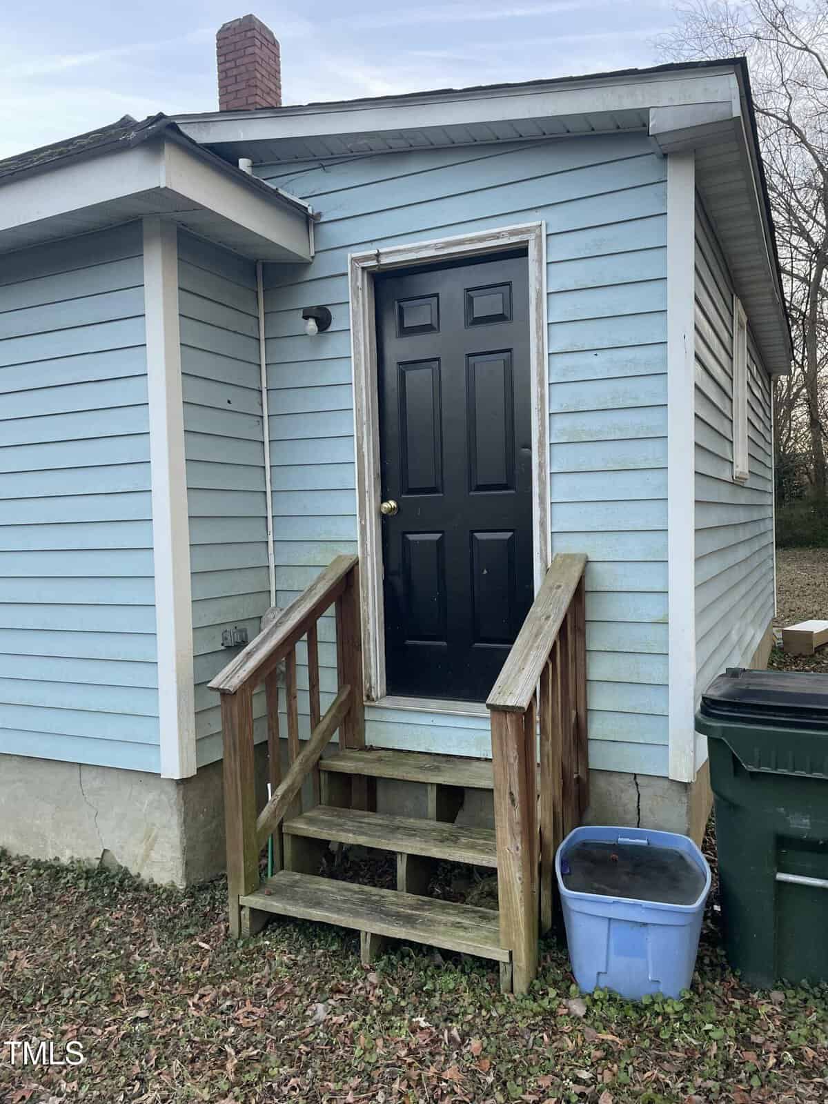 A small, light blue house with a black door and a single step wooden porch. A chimney is visible on the roof. A blue plastic container and a green trash bin sit to the right of the entrance. The ground is covered with dry leaves.