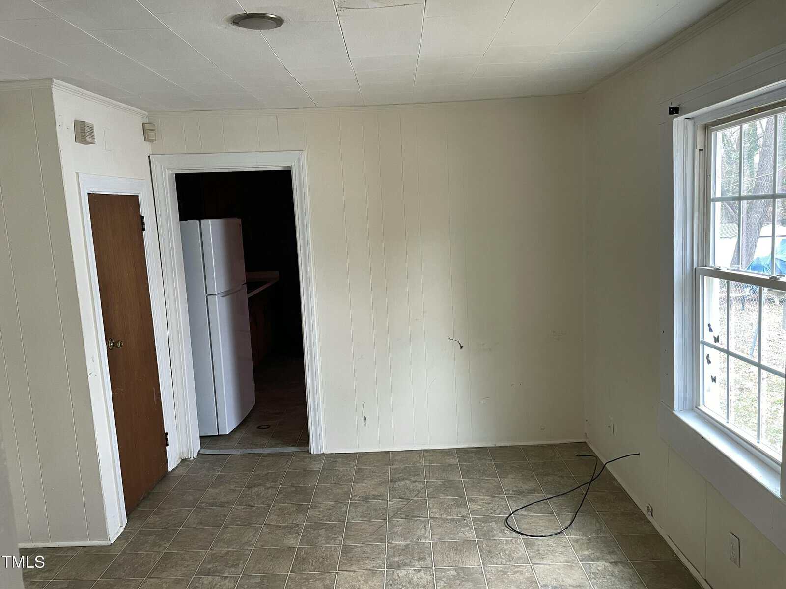An empty room with light-colored walls and a large window on the right. A wooden door and an open doorway revealing a refrigerator are on the left. The floor is covered with patterned linoleum, and a cable is coiled on the floor.