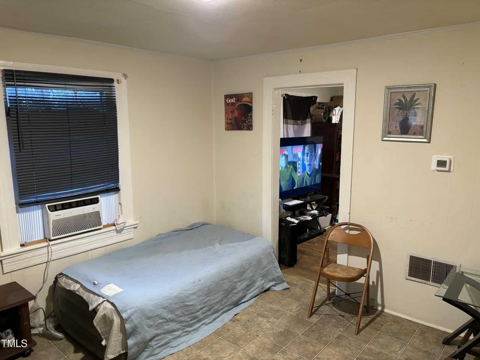 Room with a bed covered by a blue blanket, a window with a black blind, and an air conditioner. A small TV shows a scene with people. A wooden chair is against the wall, and a small table is beside the bed. Walls have a few pictures.