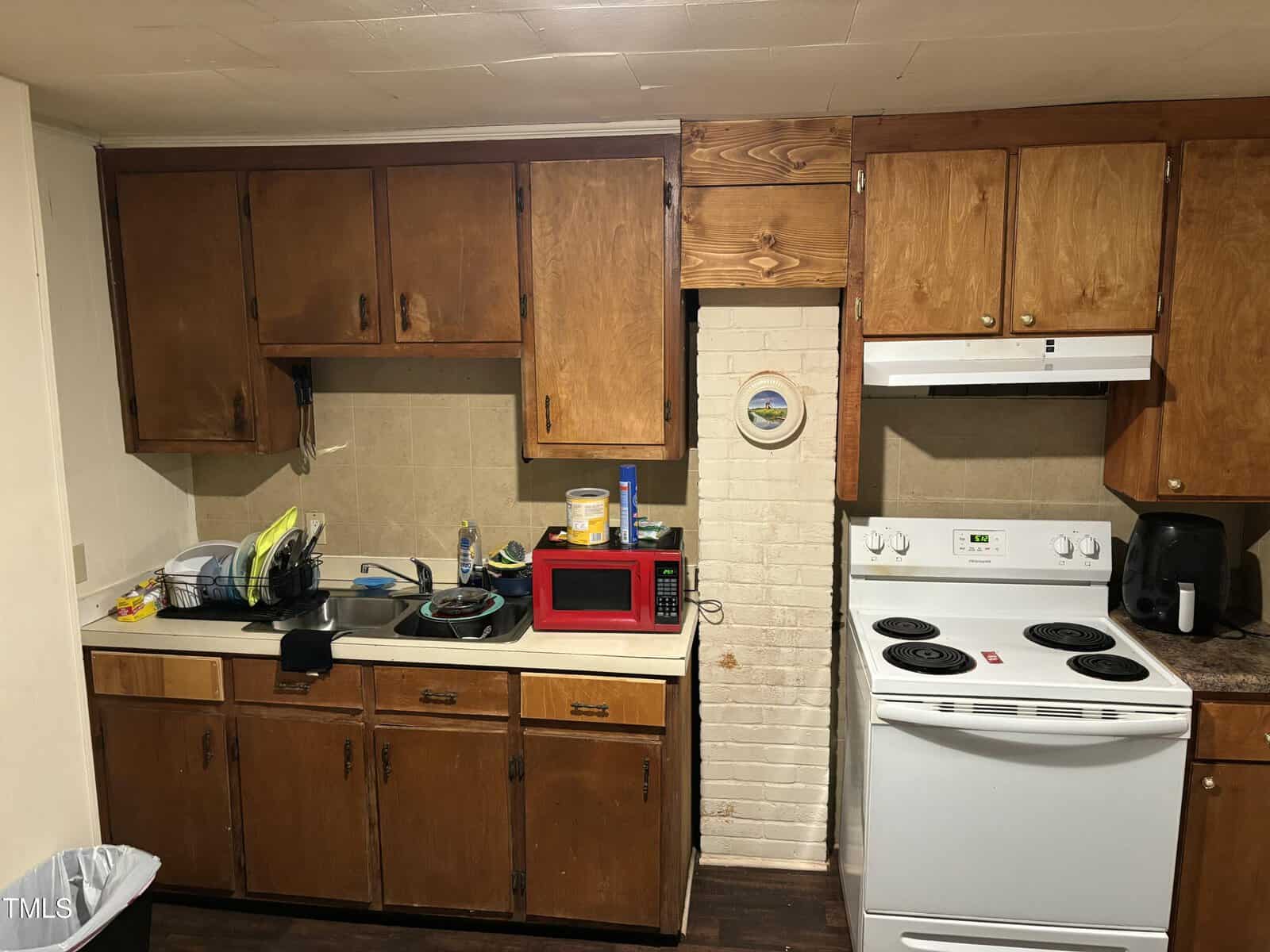 A kitchen with wooden cabinets, a white brick backsplash, white electric stove, red microwave, and countertop with a dish rack and coffee maker. Pots and cleaning supplies are visible on the counter. The floor is dark wood.