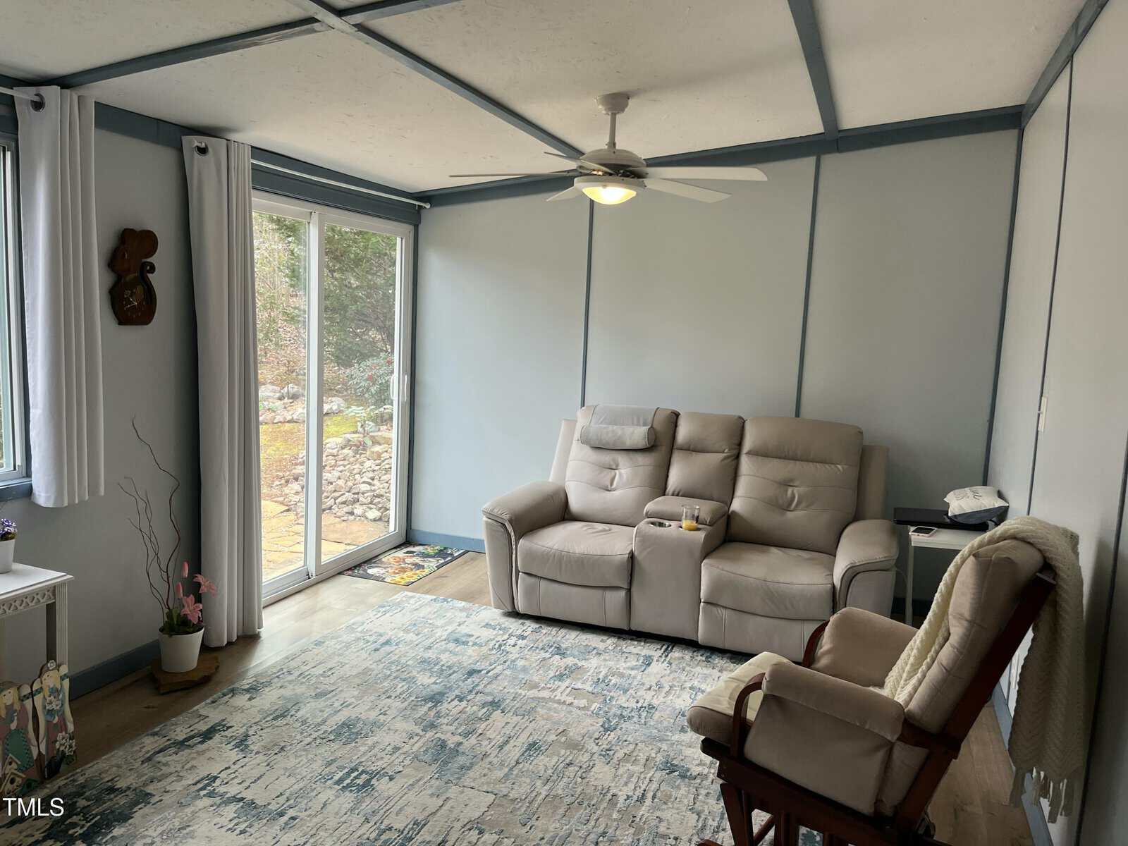 A small living room with light blue walls, a ceiling fan, a beige reclining sofa, a wooden armchair, a white cabinet, and sliding glass doors leading outside. A patterned rug covers part of the wooden floor.