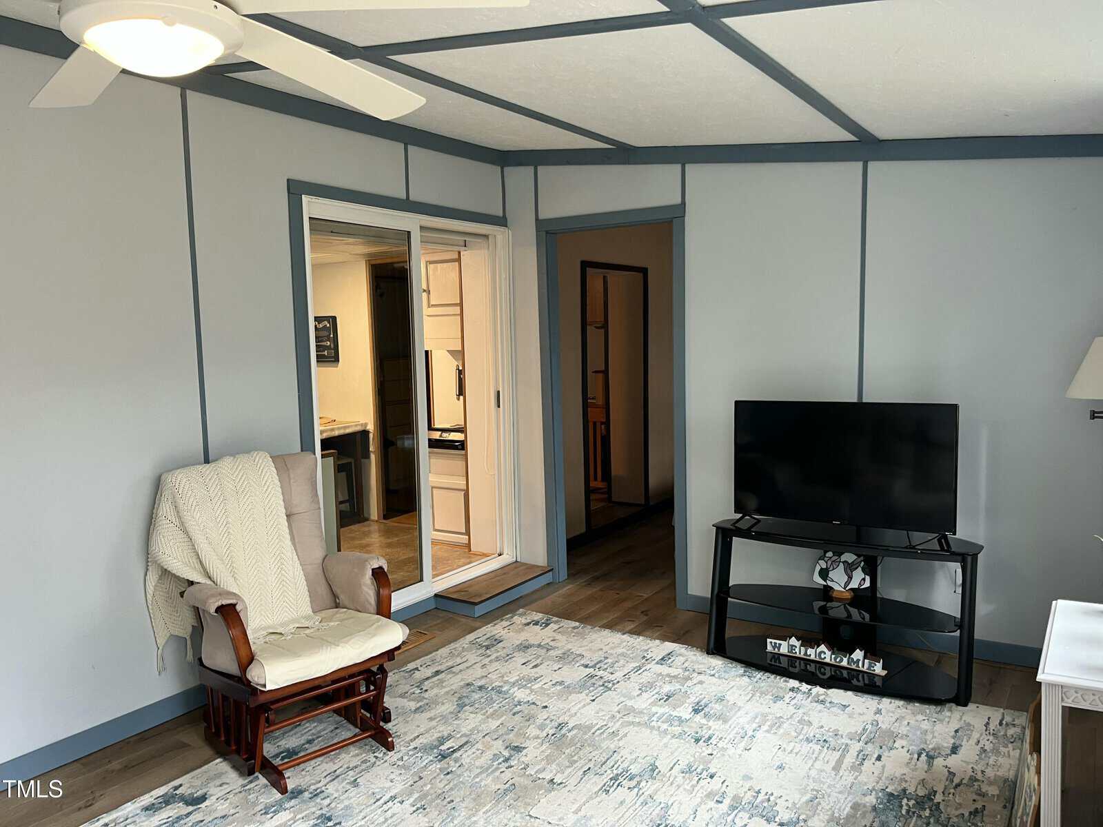 A living room with pale blue walls, a ceiling fan, a beige armchair with a blanket, a TV on a stand, and a light blue patterned rug. There is an open doorway and a sliding door leading to another room.