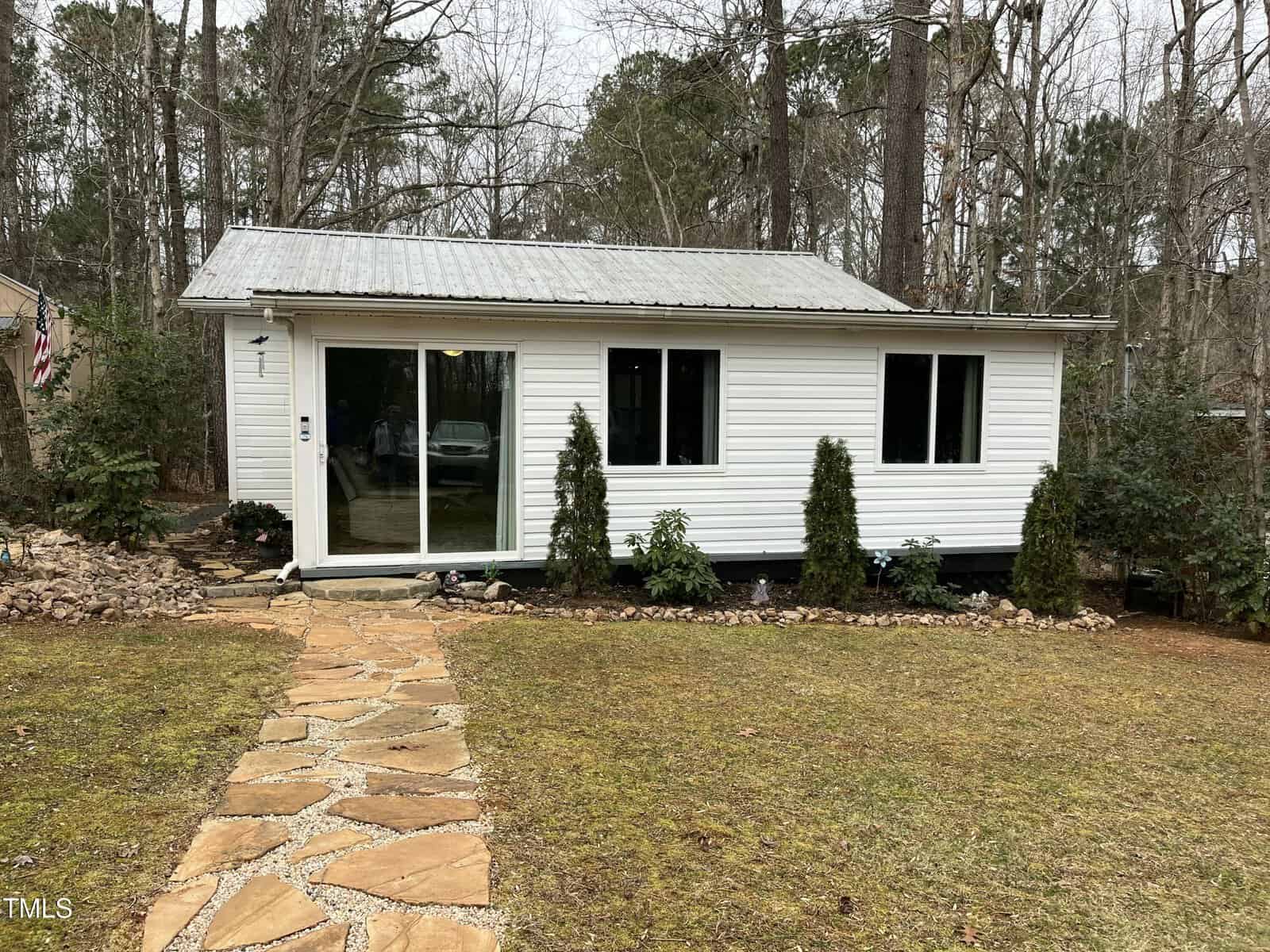 Charming small white house with a metal roof, three front windows, and a sliding glass door located at 117 Lone Star Drive in Louisburg. Shrubs line the front, with a stone path leading to the entrance. Trees and other greenery create a peaceful backdrop.