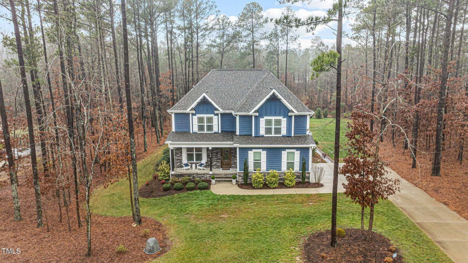 Two-story blue house with white trim, front porch, and attached garage, surrounded by tall trees and brown autumn leaves. Concrete driveway leads to the house, and the lawn is neatly landscaped.