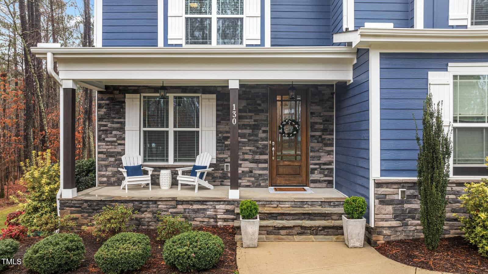 A front porch of a blue house with stone accents, two white Adirondack chairs, a small round table, potted shrubs, and a wooden front door with a wreath. Steps lead up to the porch, and landscaped plants border the walkway.