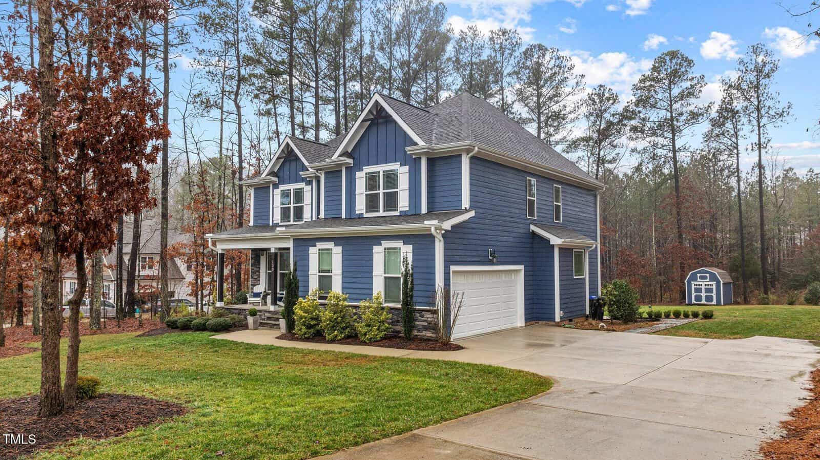 A two-story blue house with white trim, a covered front porch, attached two-car garage, and a manicured lawn. Tall trees surround the property, and a small blue shed sits in the background.
