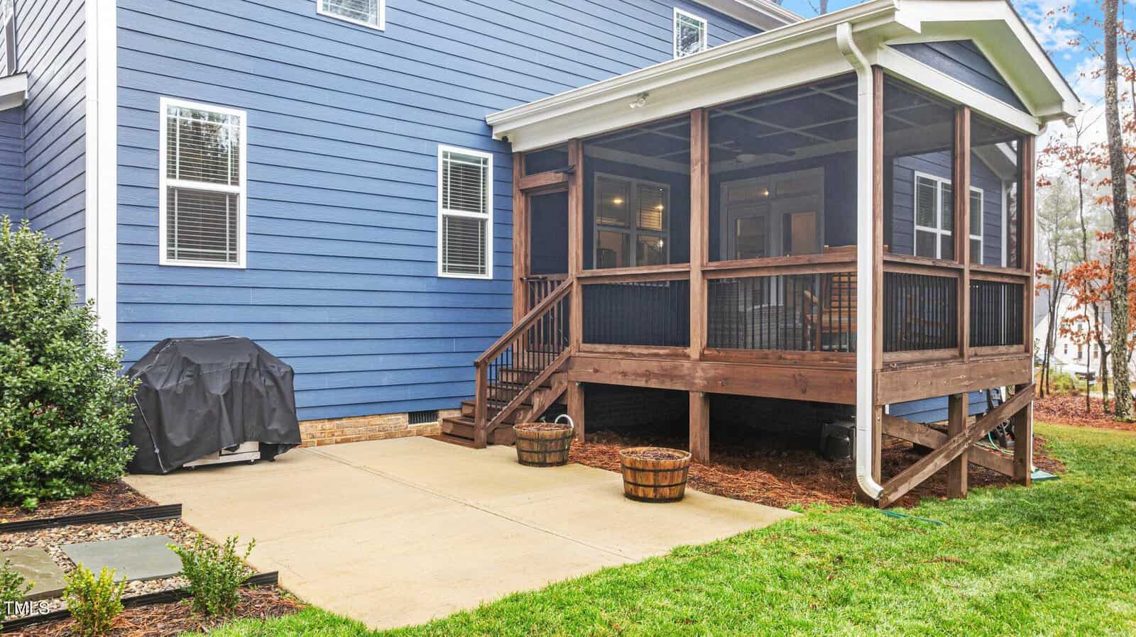A blue house with white trim features a small screened porch with wooden steps leading to a concrete patio. A covered grill and two empty wooden planters are on the patio. Green grass and small plants border the area.