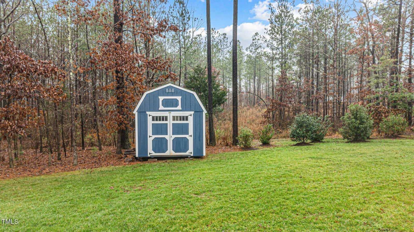 A small blue and white shed sits on green grass near the edge of a wooded area with tall trees and shrubs under a partly cloudy sky.