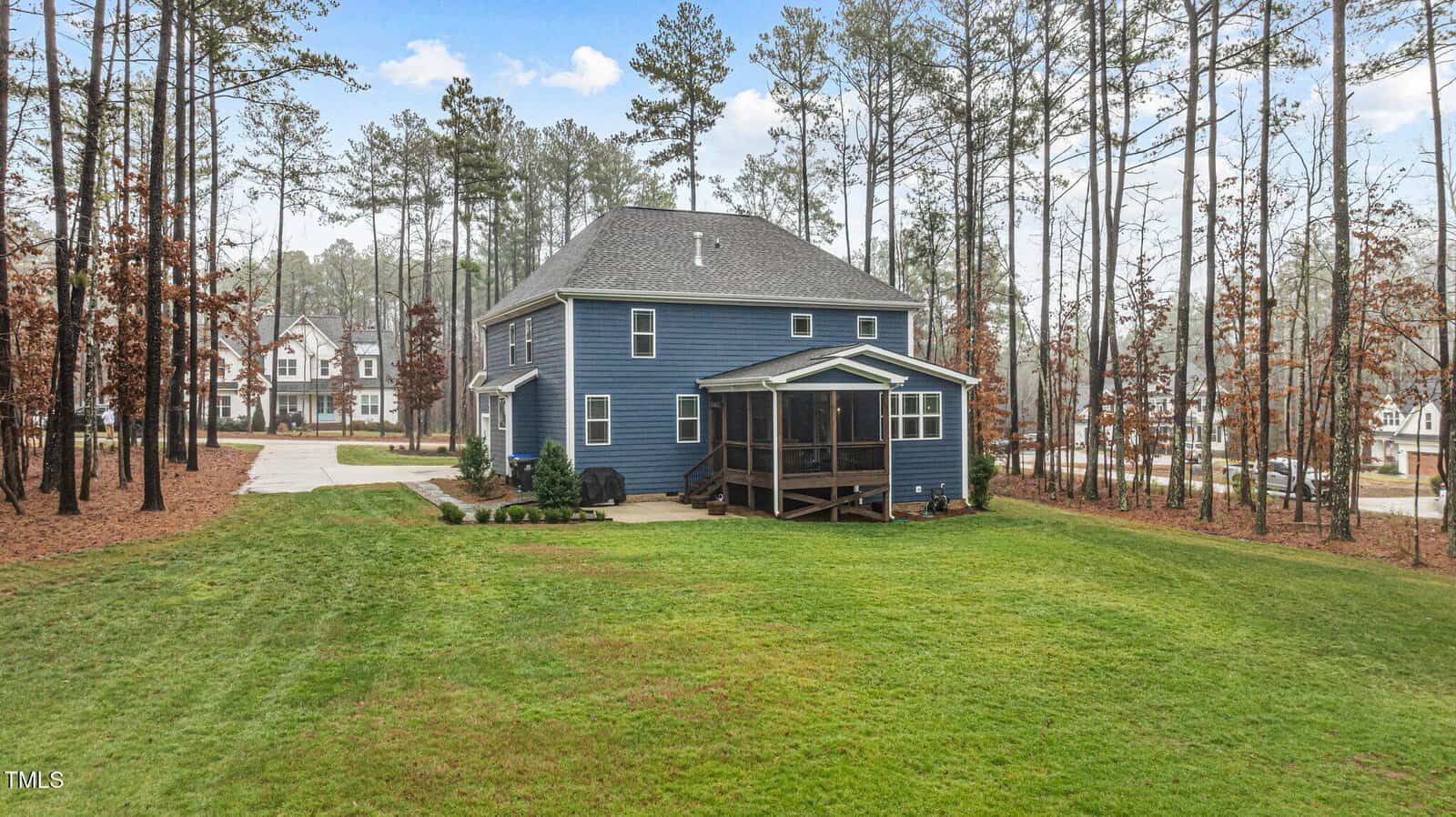 A two-story blue house with a screened porch sits on a spacious, grassy lot surrounded by tall trees. A driveway extends from the side, and neighboring houses are visible in the background.