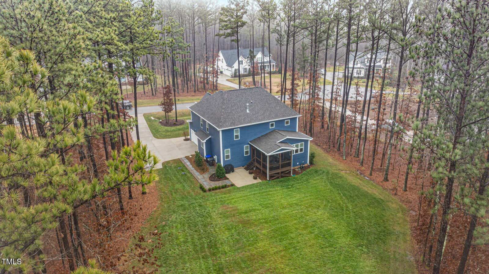A two-story blue house with a screened porch sits on a large grassy lot surrounded by tall pine trees in a suburban neighborhood, with other homes and a street visible in the background.