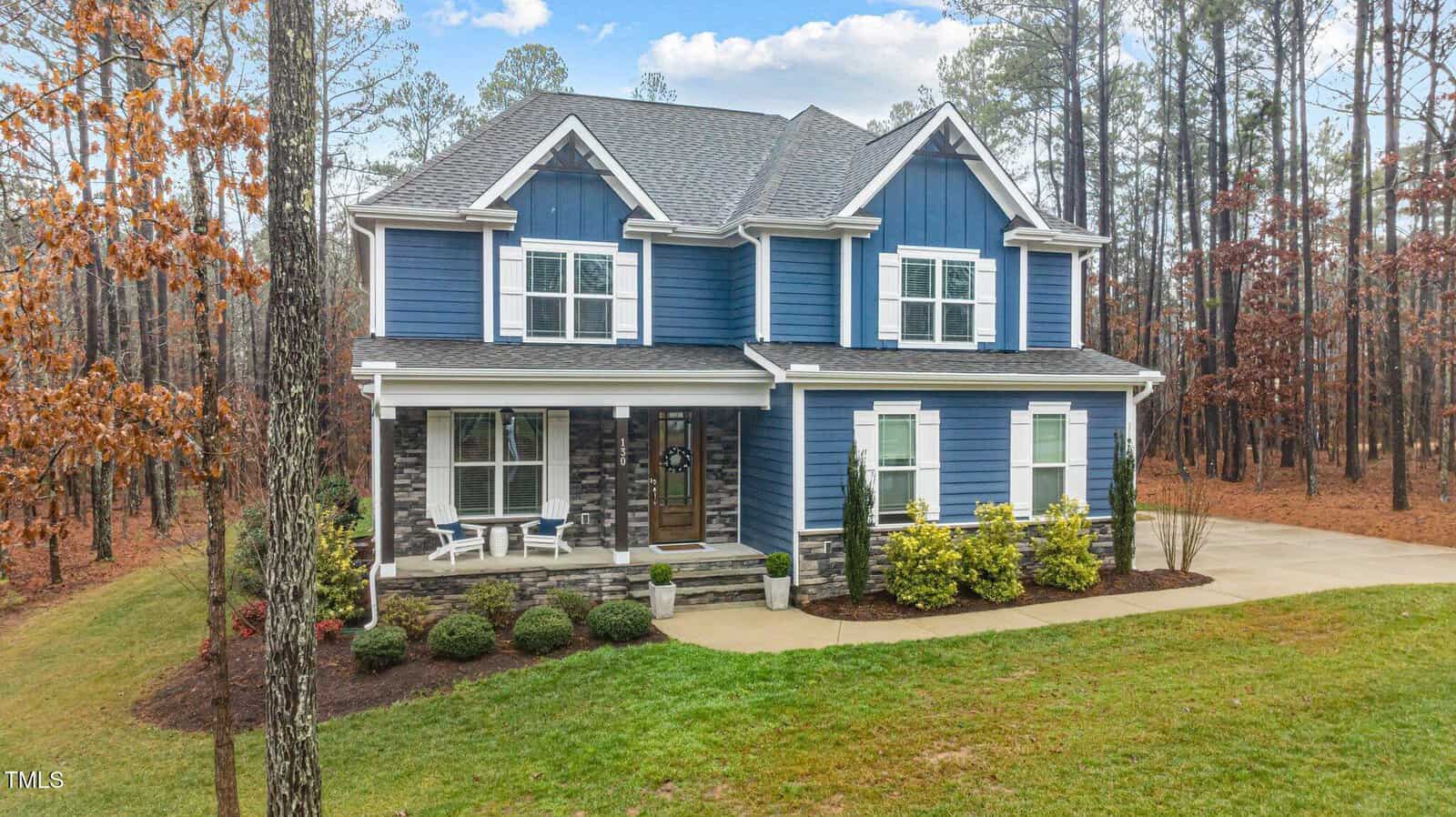 A two-story blue house with white trim, stone accents on the lower facade, a covered front porch, and a curved driveway, surrounded by trees and a well-maintained lawn.