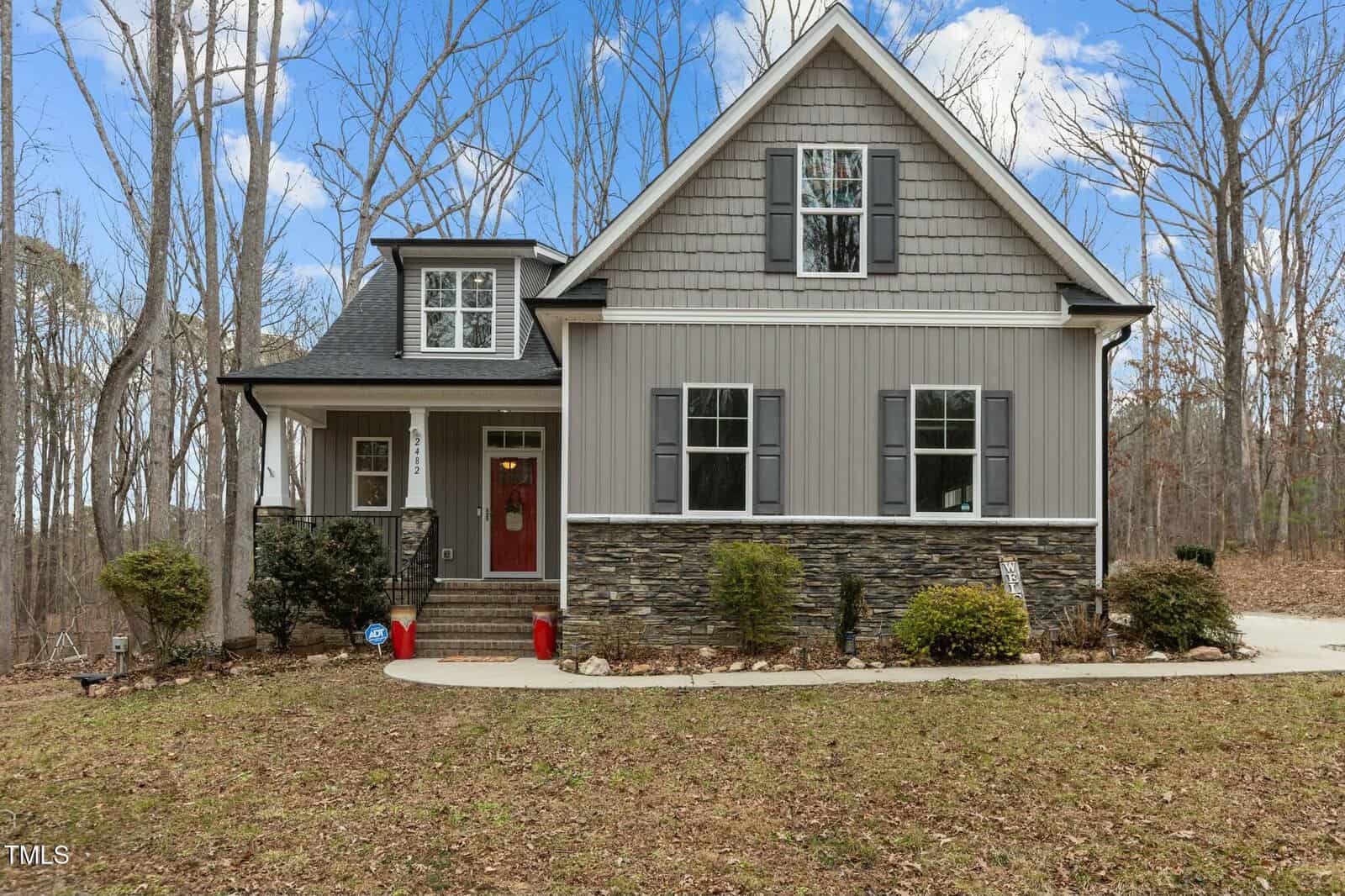 Nestled in a wooded area, the two-story house at 2482 Golden Forest Drive in Franklinton boasts gray siding, stone accents, and white trim. A charming front porch with white railings and a red door invites visitors up the steps, flanked by trees and a convenient driveway to the right.