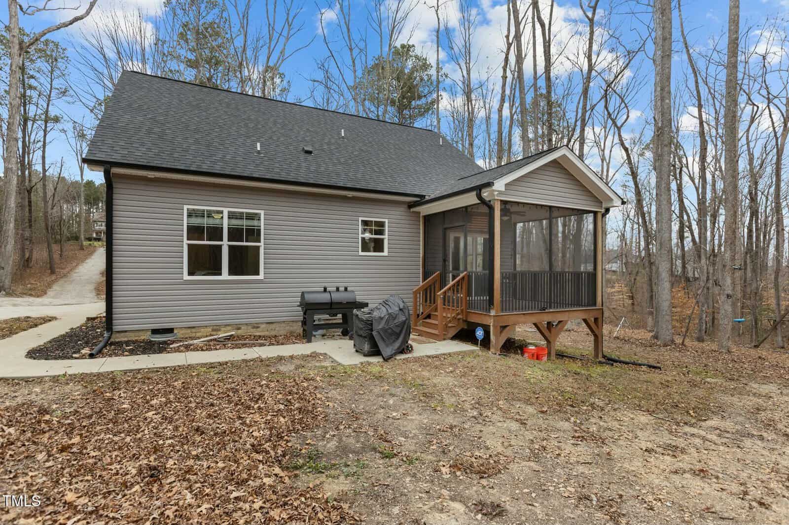 A small house with gray siding and a dark roof is situated in a wooded area. It features a screened porch with wooden steps. A black grill and a covered item are near the porch. The ground is covered with dry leaves. A concrete path leads to the house.