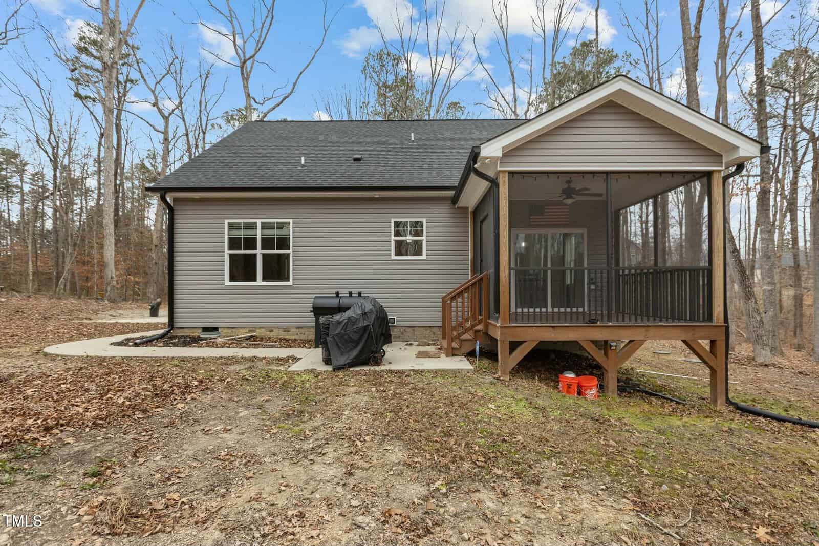 A small house with gray siding and a black roof is surrounded by a wooded area. The back features a screened porch and a grill. The yard is covered with dry leaves and patches of grass. The sky is partly cloudy.