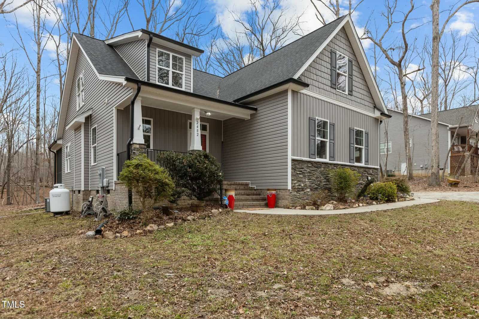 A two-story house with gray siding and stone accents is surrounded by leafless trees. It has a covered porch, multiple windows, and a dark roof. Three red planters are placed beside the staircase leading to the front door. The sky is partly cloudy.