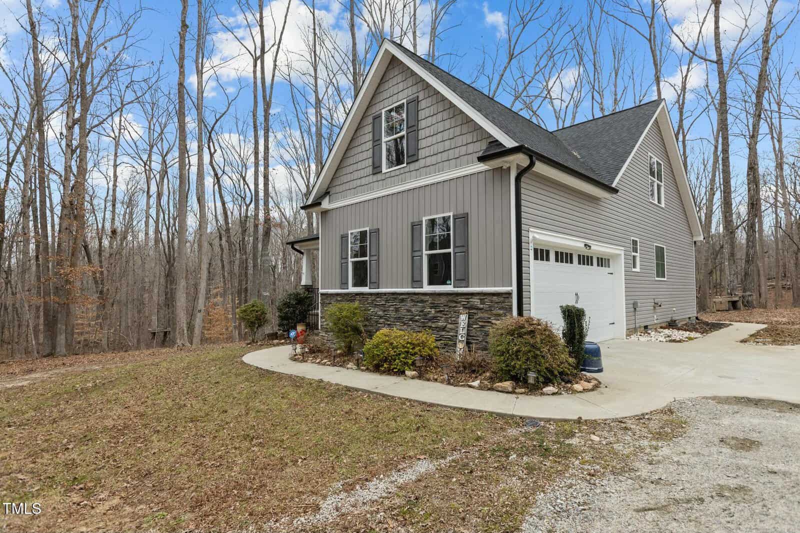 A modern, two-story house with gray siding and a stone facade is shown. It has a white garage door and is surrounded by a wooded area with bare trees. The driveway is paved, and a sidewalk leads to the entrance. The sky is partly cloudy.