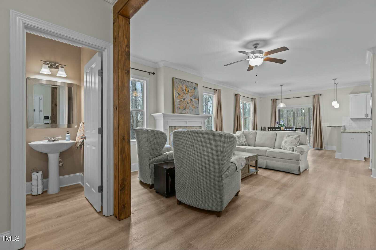 Spacious living area with light wood flooring, featuring a beige sofa and two armchairs facing a fireplace. A ceiling fan and recessed lighting illuminate the room. An open doorway leads to a small, neatly arranged bathroom.