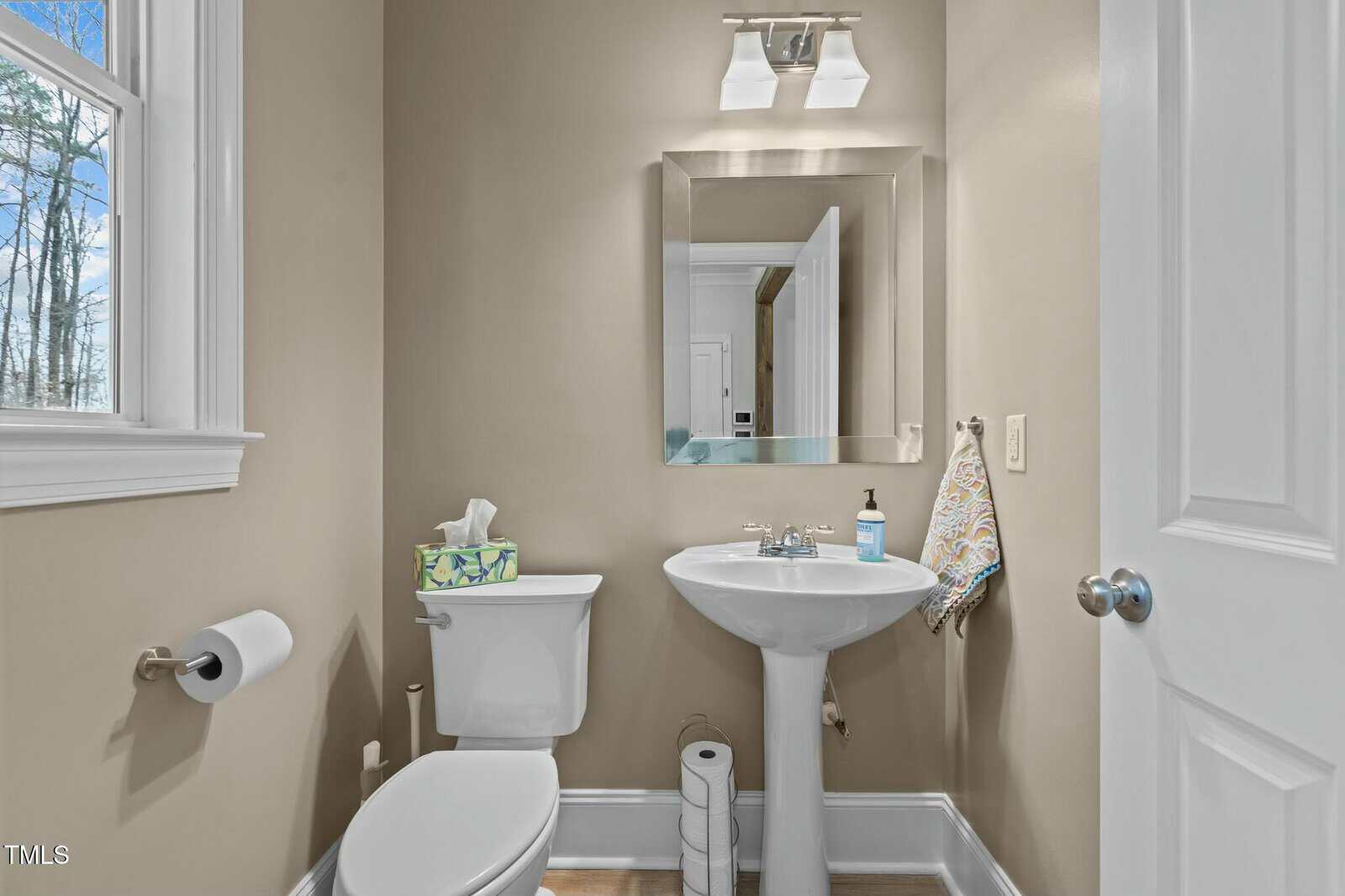 A small bathroom with a pedestal sink and a toilet. Above the sink is a square mirror and a wall-mounted light fixture. A hand towel hangs on a ring, and a window on the left allows natural light. A tissue box and soap dispenser sit on the sink.