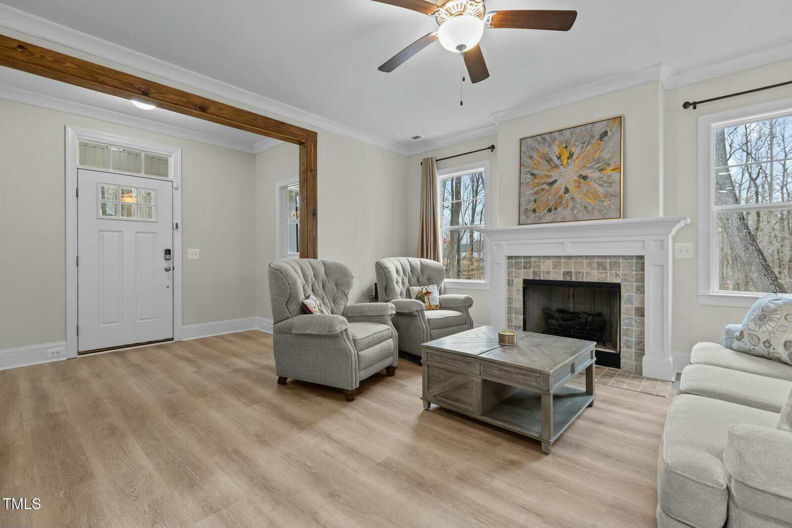 A living room with light wood flooring, featuring a ceiling fan. It has two armchairs and a sofa, a large coffee table, and a fireplace with artwork above it. Two windows on either side of the fireplace allow natural light into the room.