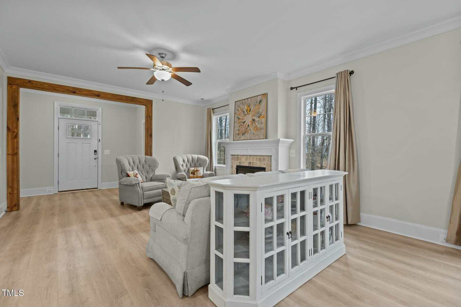 A living room with light wooden floors, featuring two armchairs near a white fireplace. A wooden beam frames the entrance, and a ceiling fan is above. There's a glass-fronted cabinet and two windows with drapes.