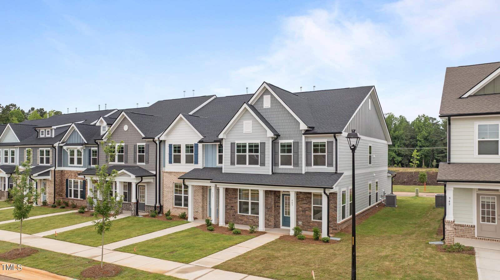 A row of modern, two-story townhouses with gray siding, white trim, and brick accents, each with small front yards and driveways, is seen under a clear blue sky.