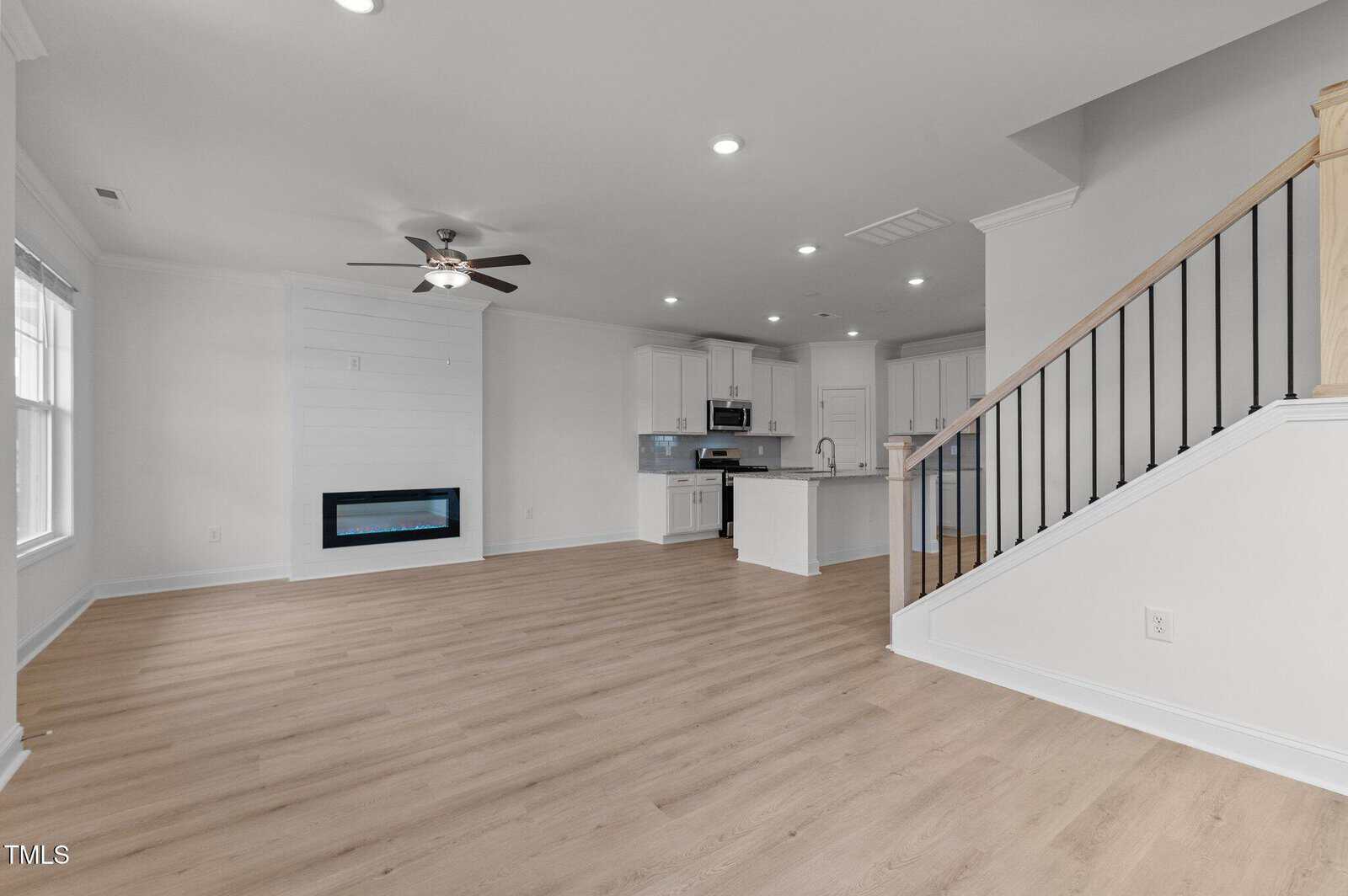 Open-concept living area with light wood flooring, white walls, a ceiling fan, modern fireplace, large windows, and a kitchen with white cabinets and island. Wooden stairs with black railings are on the right.