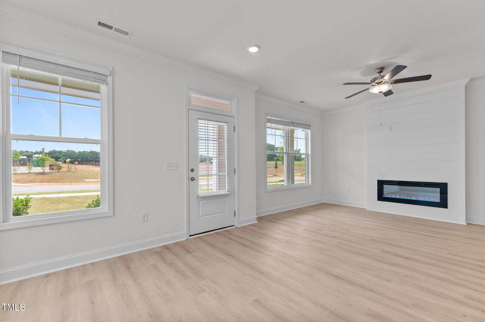 Bright, empty living room with light wood floors, white walls, large windows, a glass door, ceiling fan, and a modern built-in electric fireplace along one wall.
