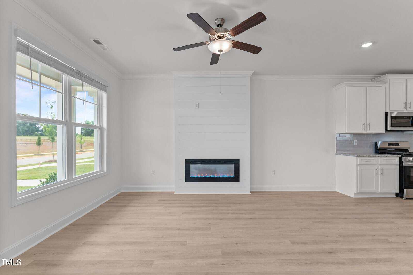 Bright, modern living room with large windows, light wood floors, white walls, ceiling fan, built-in electric fireplace, and part of a white kitchen with stainless steel appliances visible on the right.