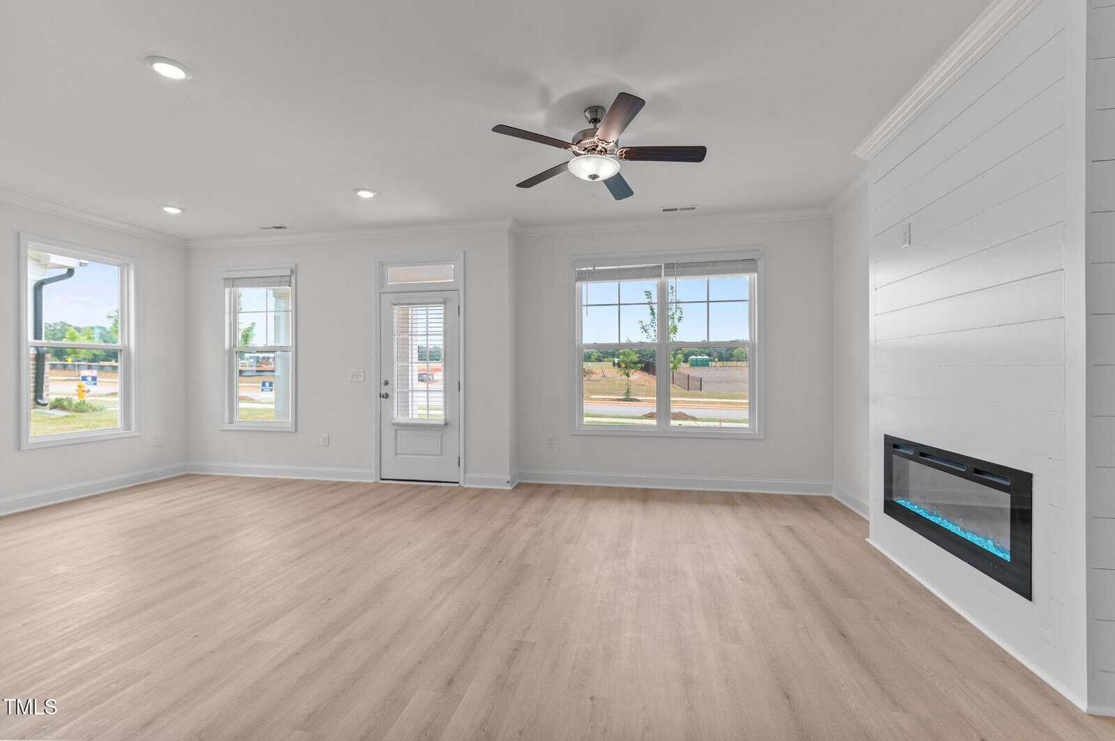 Bright, empty living room with large windows, light wood floors, white walls, a ceiling fan, wall-mounted electric fireplace, and a glass door leading outside. Ample natural light fills the space.