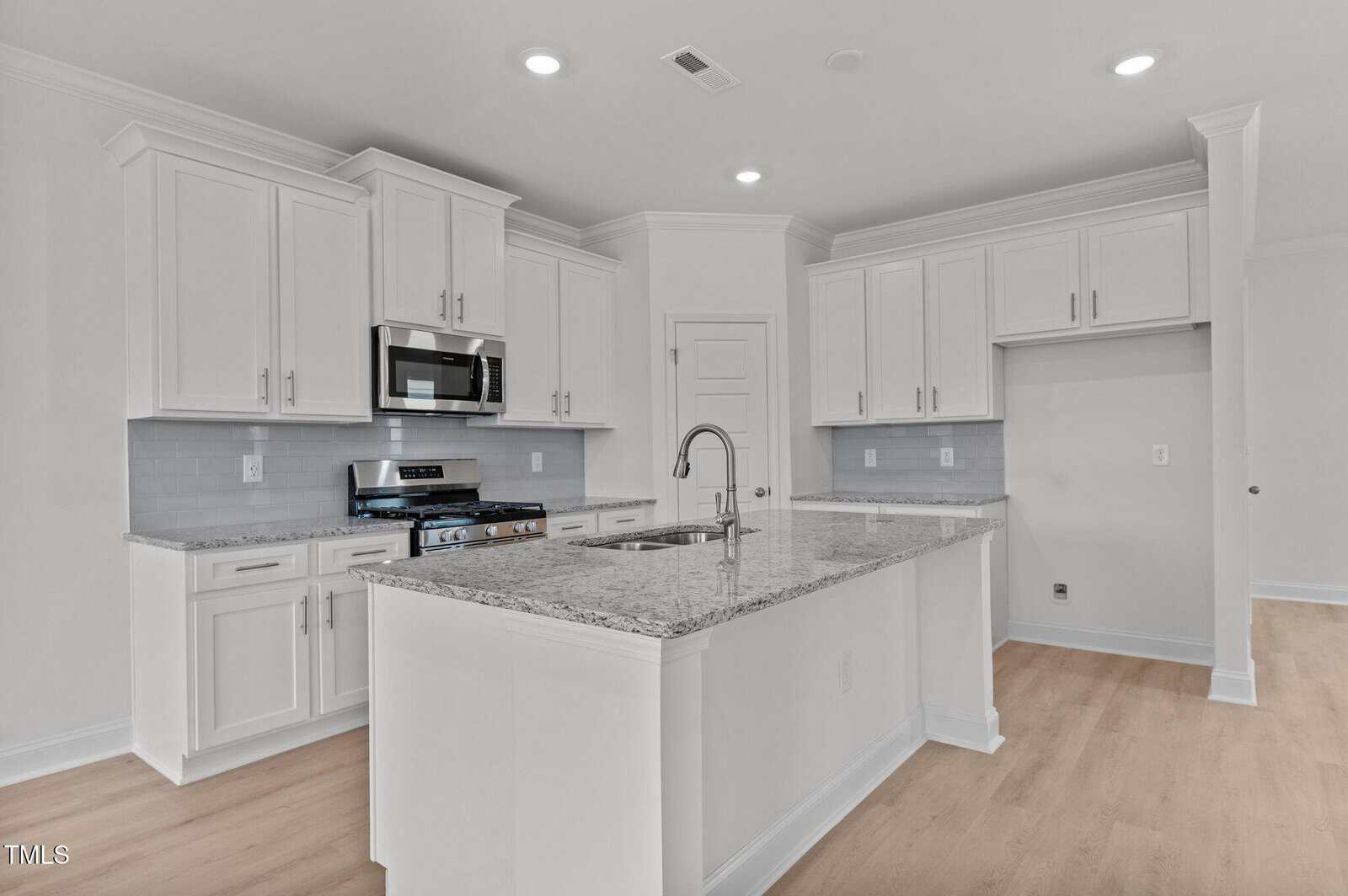 Modern kitchen with white cabinets, stainless steel appliances, a granite-topped island with a built-in sink, light wood flooring, and recessed ceiling lights. The space is bright and uncluttered.