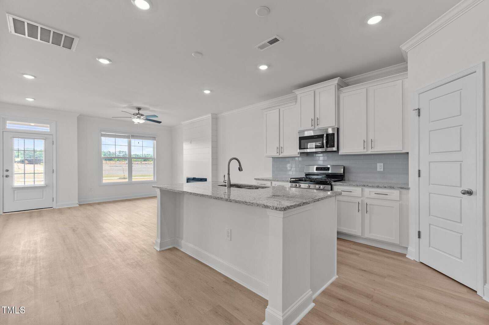 Modern kitchen with white cabinets, stainless steel appliances, a large center island with granite countertop, and wood floors. Open layout connects to a bright living area with large windows and a door to the outside.