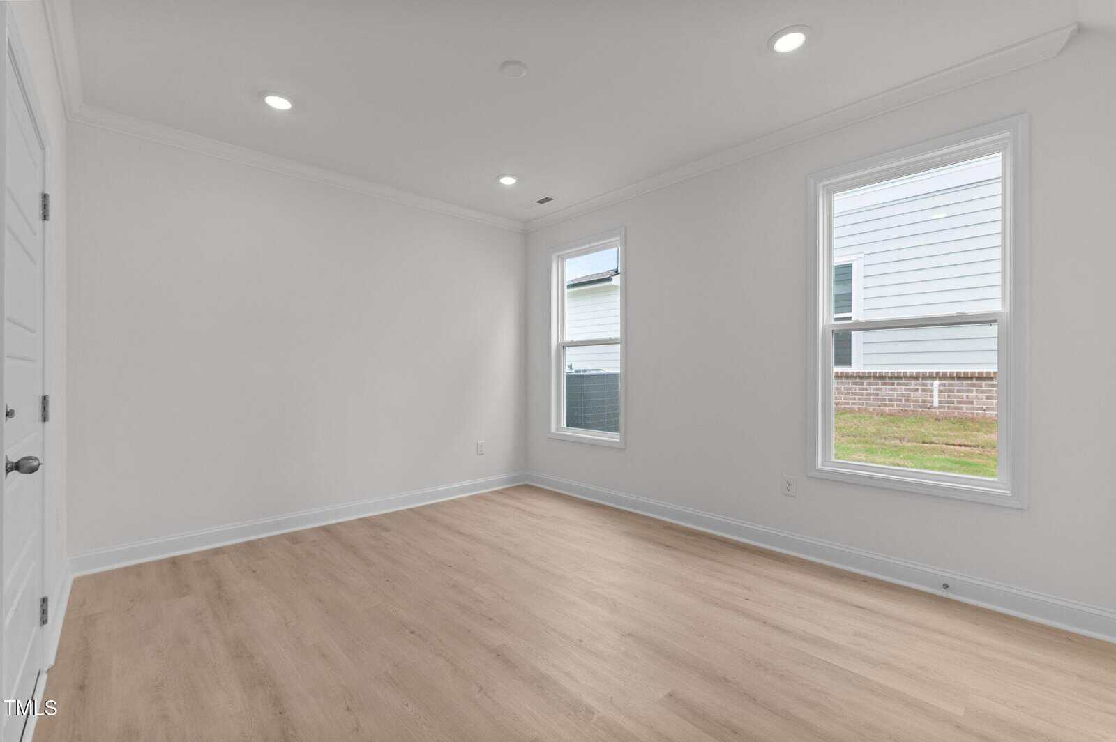 A bright, empty room with white walls, two large windows, light wood flooring, recessed ceiling lights, and a white door. Sunlight comes through the windows, showing a view of a fenced yard and another building outside.