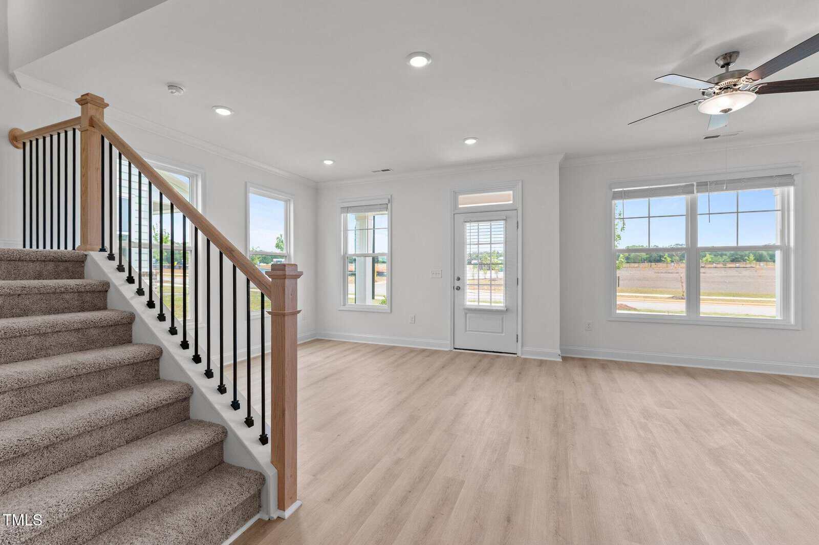 Bright, empty living room with light wood floors, white walls, several windows, a ceiling fan, and a carpeted staircase with a wooden handrail and black spindles.