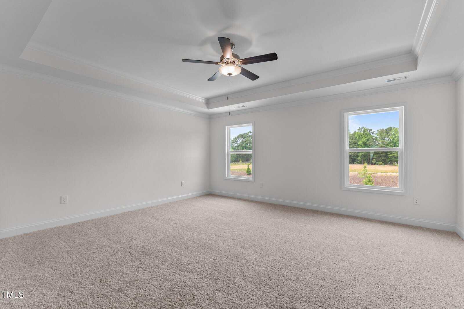 Empty, carpeted room with white walls and two large windows letting in natural light. A ceiling fan with lights is centered on the ceiling, and the room has crown molding and a tray ceiling design.