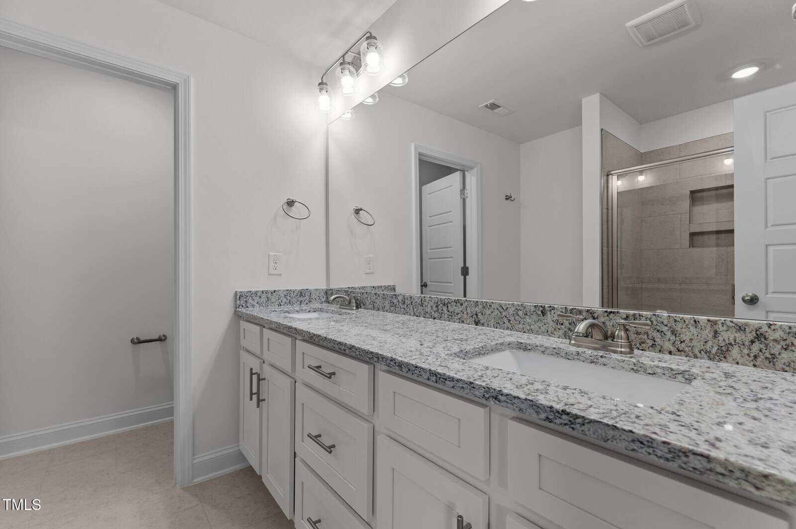 Modern bathroom with dual sinks, granite countertop, white cabinets, large wall mirror, and bright lighting. A shower with glass doors is visible in the background. Walls and doors are painted white.