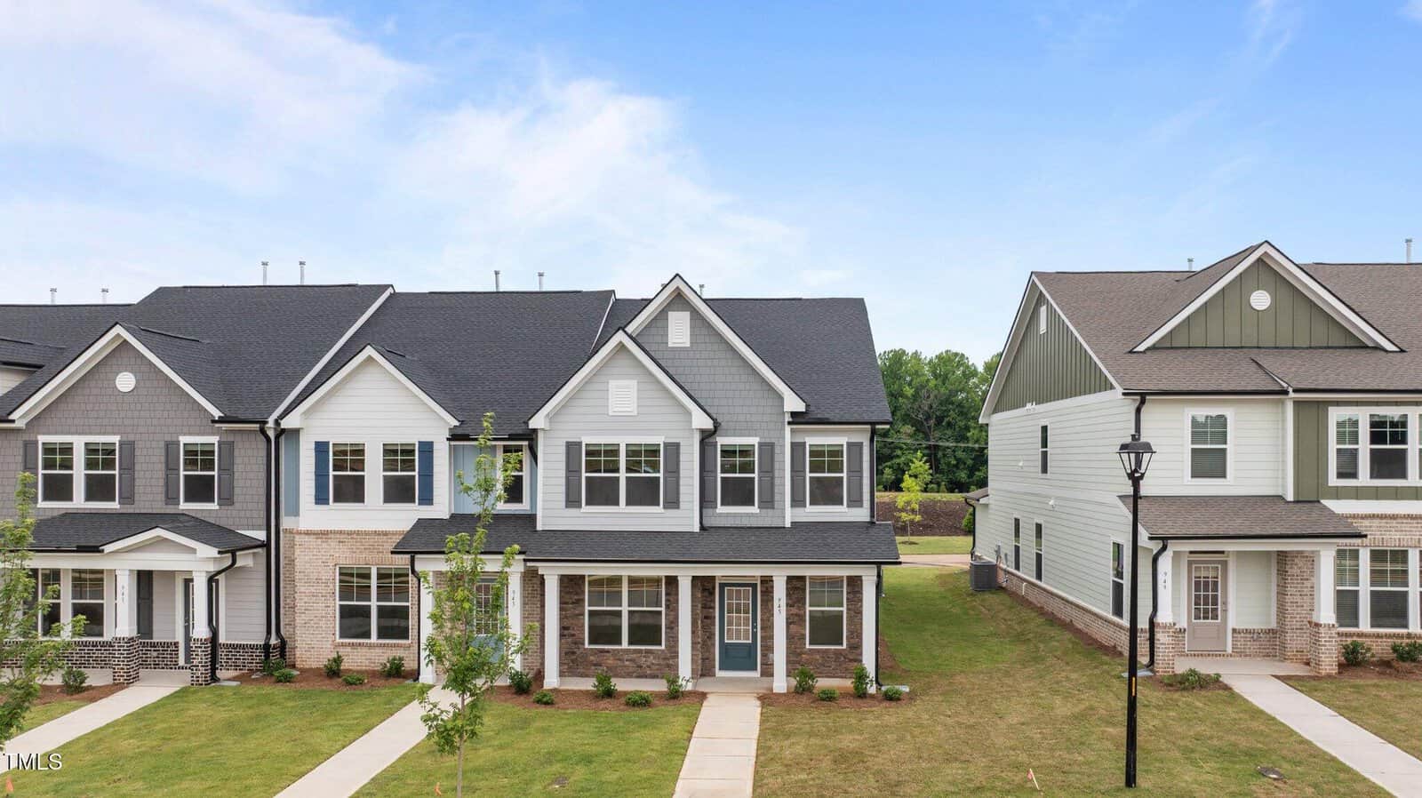 A row of modern two-story townhouses with white and gray exteriors, front porches, manicured lawns, and a clear blue sky overhead. A sidewalk and young trees line the front.