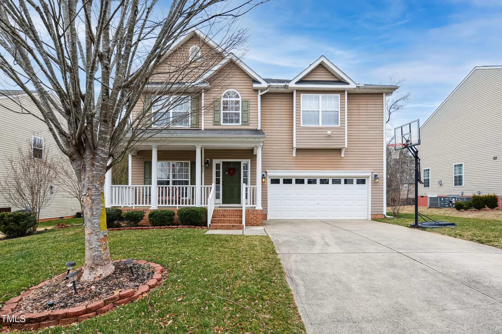 Two-story beige house with a covered front porch, white trim, attached two-car garage, and a basketball hoop by the driveway. There is a large tree and brick-edged flower bed in the front yard.