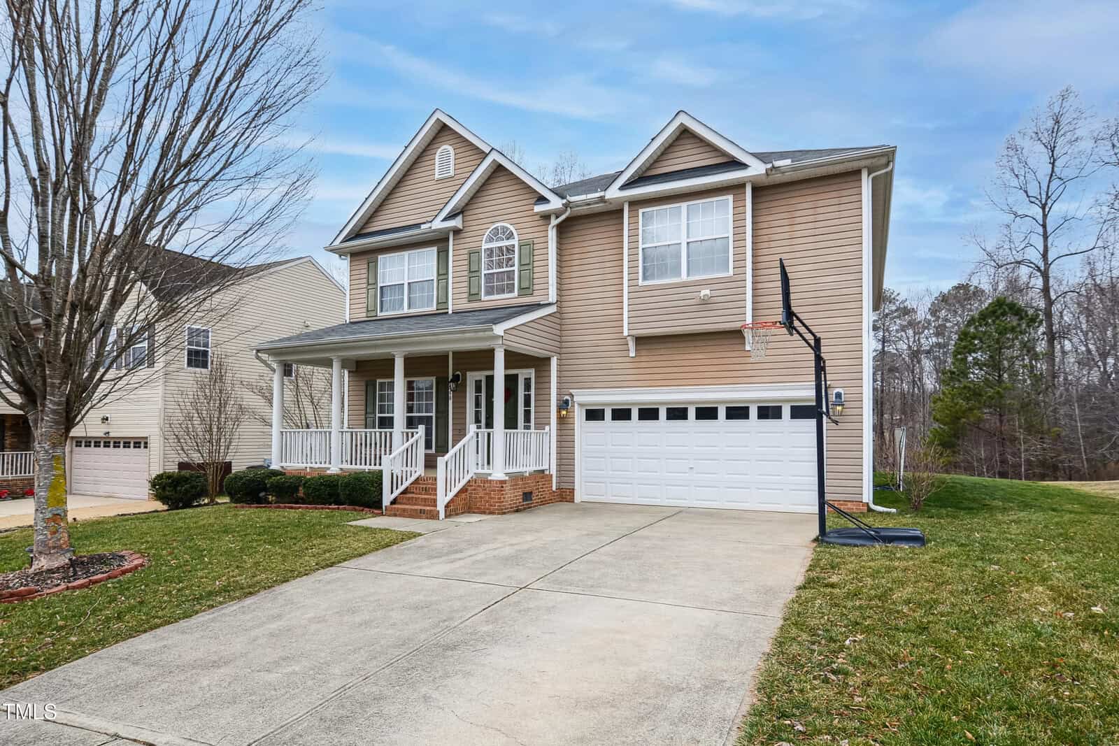 Two-story beige suburban house with white trim, a front porch, attached two-car garage, and a basketball hoop next to the driveway. Leafless tree in front yard, grass and neighboring houses visible.