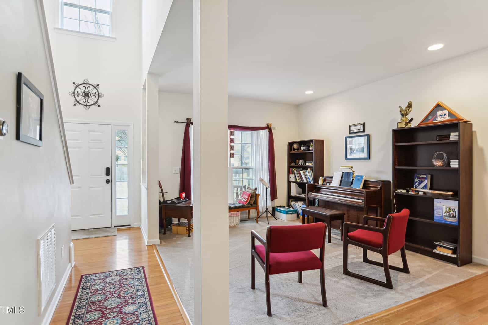 A bright living room with a piano, two red chairs, bookshelves, and framed pictures. The front door and a hallway with a rug are visible, along with large windows letting in natural light.