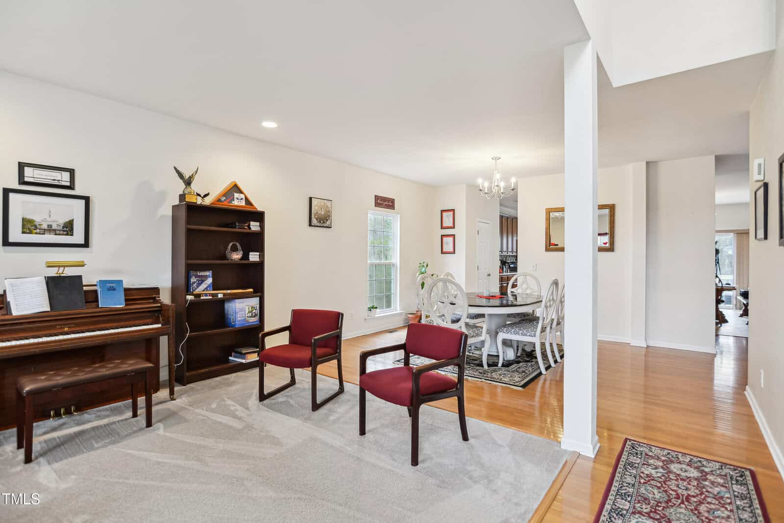 A living room with a piano, bookshelf, two red chairs, and a beige carpet, adjacent to a dining area with a round table, six chairs, chandelier, hardwood floors, and framed artwork on the walls.
