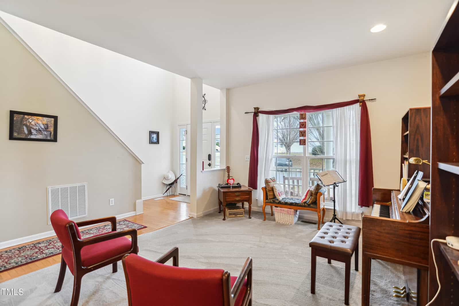 A living room with beige walls, a light carpet, red chairs, a wooden piano, a bookshelf, and large windows with red curtains. The entryway and a staircase are visible in the background.