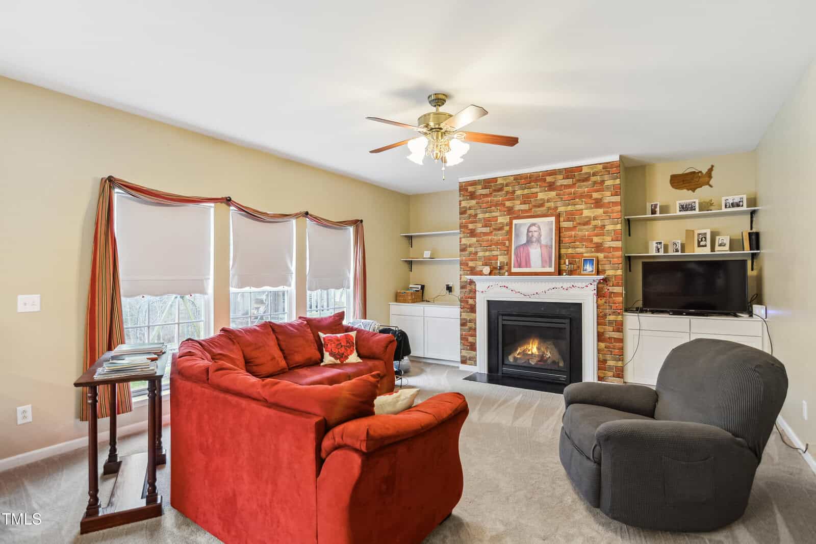 Living room with a red sectional sofa, gray armchair, stone fireplace, TV on shelves, ceiling fan, and three windows with closed blinds. A framed picture and garland are on the mantel above the fireplace.