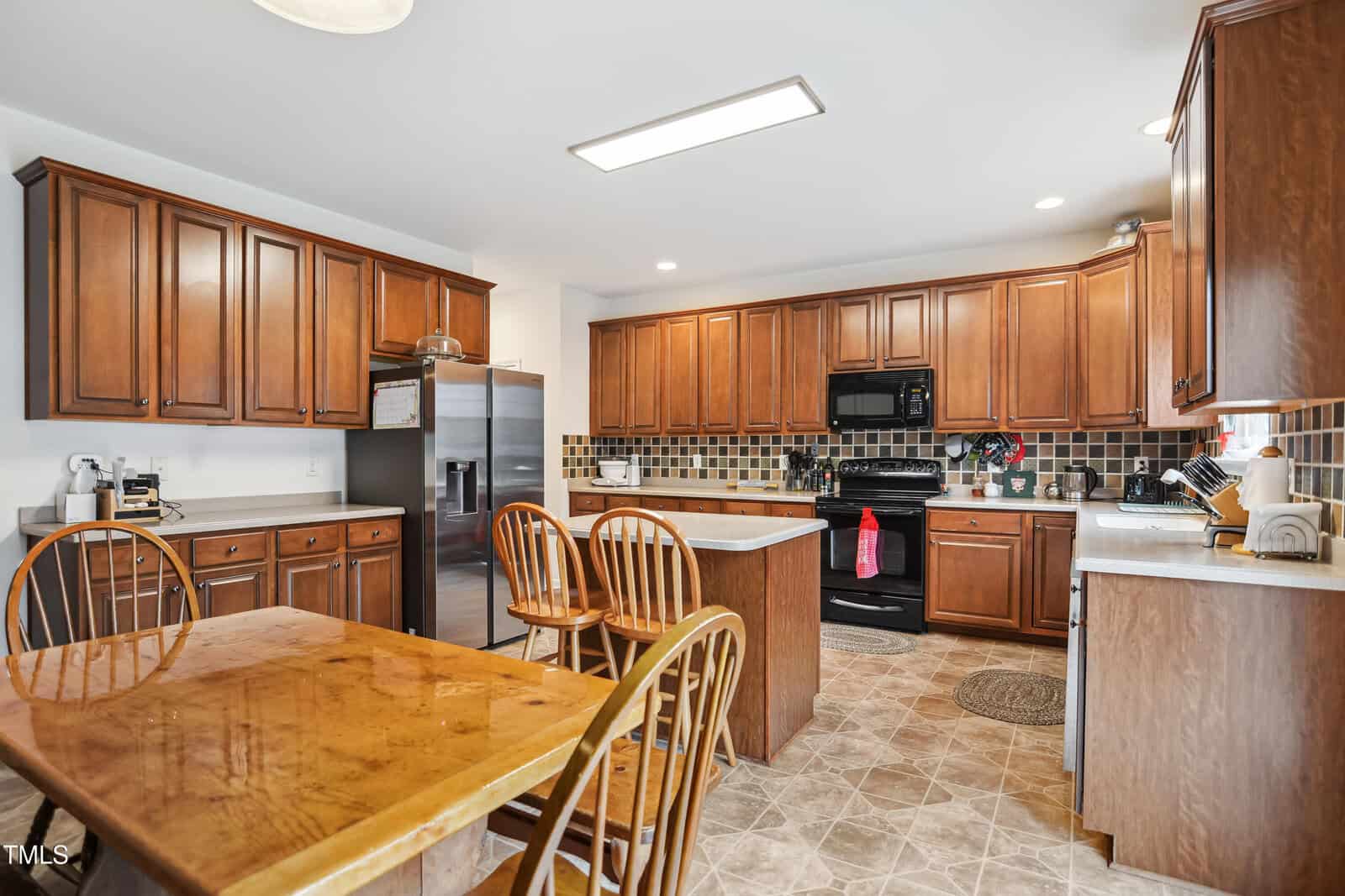 Spacious kitchen with wooden cabinets, tile backsplash, stainless steel refrigerator, black appliances, and a wooden dining table with chairs. Tiled floor and various kitchen items on counters. Bright, natural lighting.