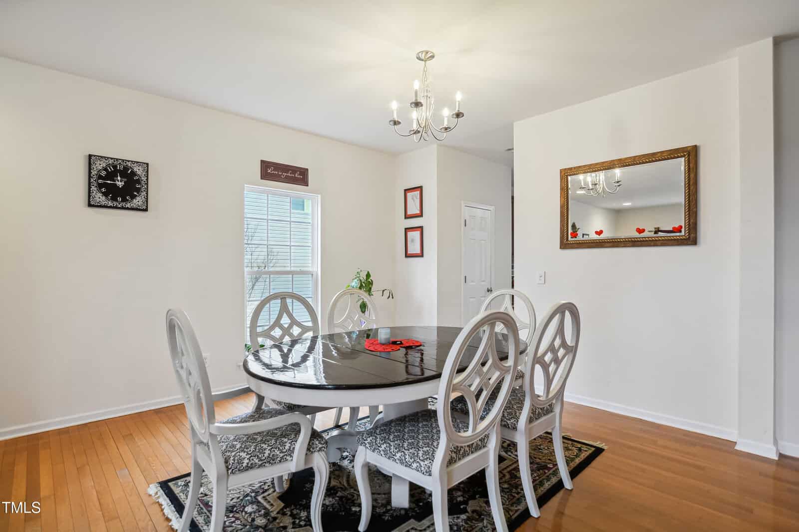 A dining room with a round table and six white chairs, a black-patterned rug, a wall mirror, chandelier, and framed wall art. Wooden floors and white walls complete the space. A window lets in natural light.